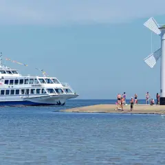 Biała latarnia morska wiatrakowa, przypominająca młyn, stoi na piaszczystym cyplu. Obok cumuje biało-niebieski statek wycieczkowy. Kilka osób odpoczywa na brzegu. To jeden z urokliwych widoków na polskim wybrzeżu.