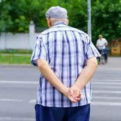 Starszy pan w kratowanej koszuli i czapce, trzymając ręce za plecami, czeka na zielone światło na pasach.
