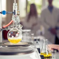 W laboratorium trwa eksperyment. W kolbie okrągłodennej, podgrzewanej na płycie, bulgocze żółty płyn. W tle widać naukowców w kitlach, badających wartościowość pierwiastków.