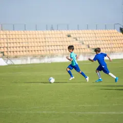 Dwóch chłopców w strojach **sporty po angielsku** biega po boisku piłkarskim, jeden z nich prowadzi piłkę.