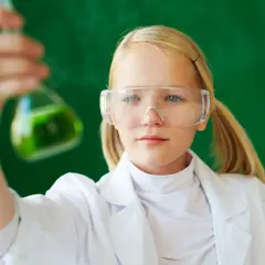 Dziewczynka w okularach ochronnych i białym fartuchu trzyma kolbę z zielonym płynem. To przykład edukacji dla bezpieczeństwa w laboratorium.