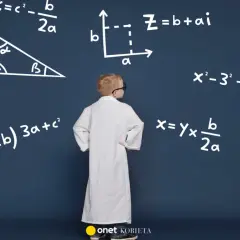 Chłopiec w białym fartuchu stoi przed tablicą z wzorami, w tym wzorem na przyspieszenie.
