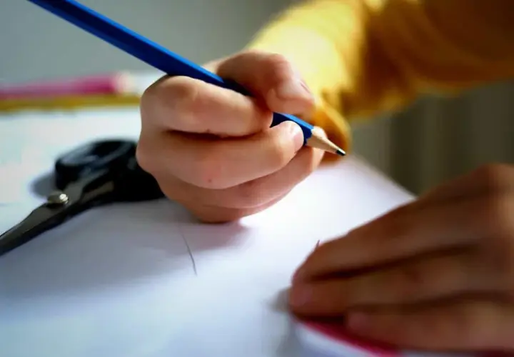 Dłoń podmiotu lirycznego trzyma niebieską kredkę, rysując na białej kartce. Obok leżą nożyczki.