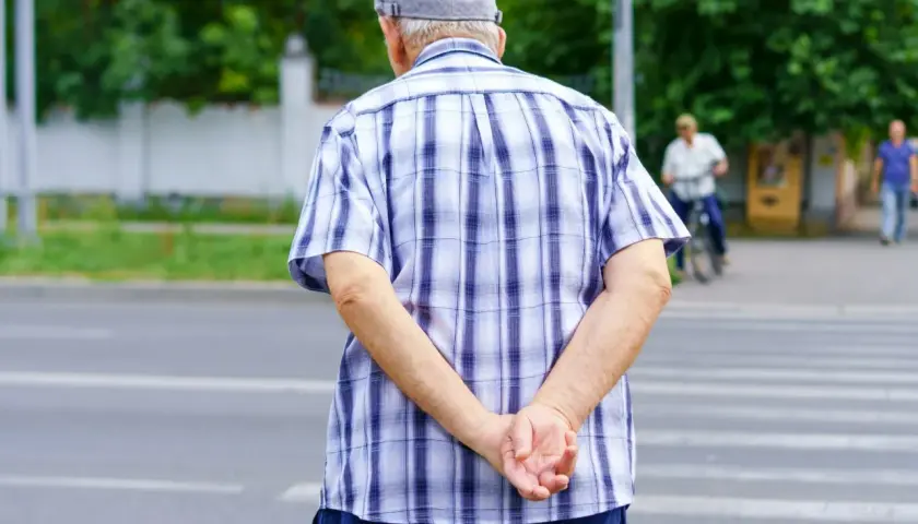 Starszy pan w kratowanej koszuli i czapce, trzymając ręce za plecami, czeka na zielone światło na pasach.