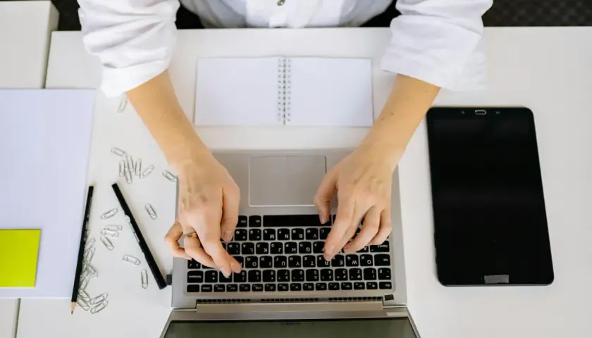 Kobieta pisze na laptopie, szukając informacji, jak napisać stopnie Celsjusza w Excelu. Obok leżą tablet, notatnik i spinacze.