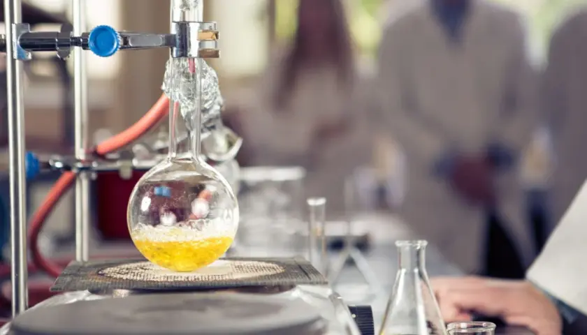 W laboratorium trwa eksperyment. W kolbie okrągłodennej, podgrzewanej na płycie, bulgocze żółty płyn. W tle widać naukowców w kitlach, badających wartościowość pierwiastków.