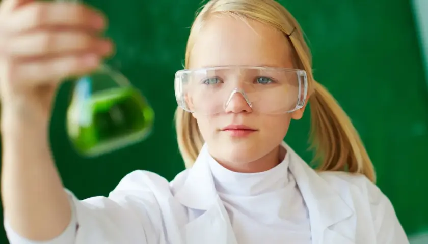 Dziewczynka w okularach ochronnych i białym fartuchu trzyma kolbę z zielonym płynem. To przykład edukacji dla bezpieczeństwa w laboratorium.