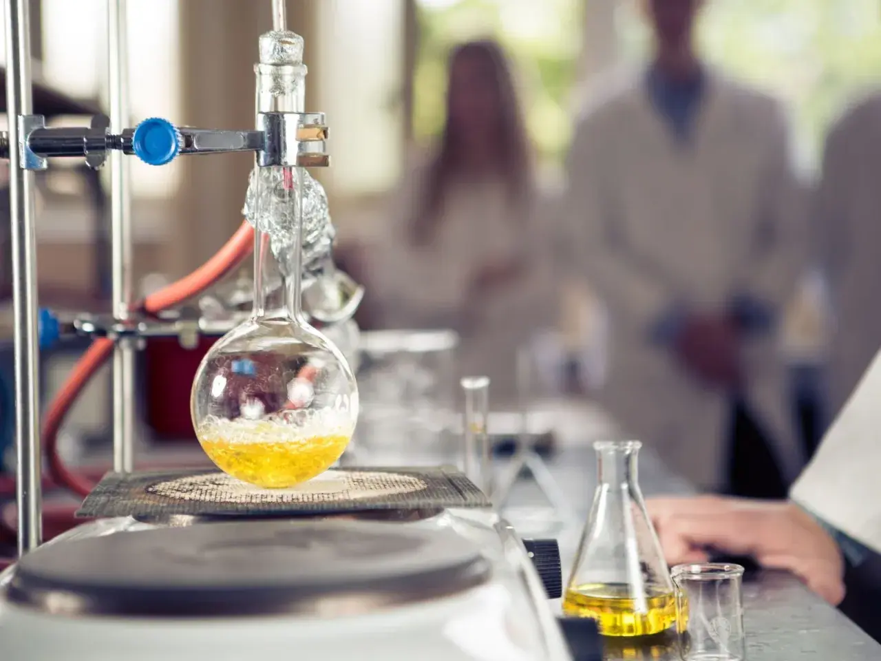 W laboratorium trwa eksperyment. W kolbie okrągłodennej, podgrzewanej na płycie, bulgocze żółty płyn. W tle widać naukowców w kitlach, badających wartościowość pierwiastków.