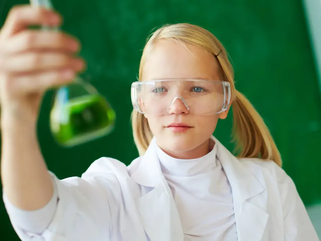 Dziewczynka w okularach ochronnych i białym fartuchu trzyma kolbę z zielonym płynem. To przykład edukacji dla bezpieczeństwa w laboratorium.